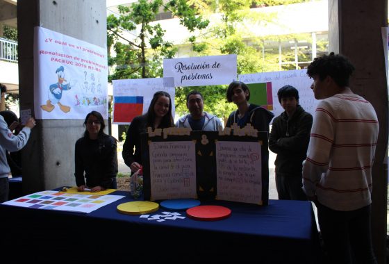 La imagen muestra un stand de la Feria de Matemática de los Talleres PACE UC realizada en el Patio de Humanidades del Campus San Joaquín en octubre de 2019. El stand denominado “Resolución de problemas” muestra al profesor Rodrigo Ramírez, acompañado de los estudiantes.