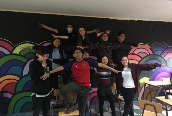 La imagen muestra a 10 estudiantes con los brazos extendidos posando delante de un mural pintado en la sala de clases de Historia del Liceo Gabriel González Videla A24 en el marco de los Proyectos Participativos 2019.