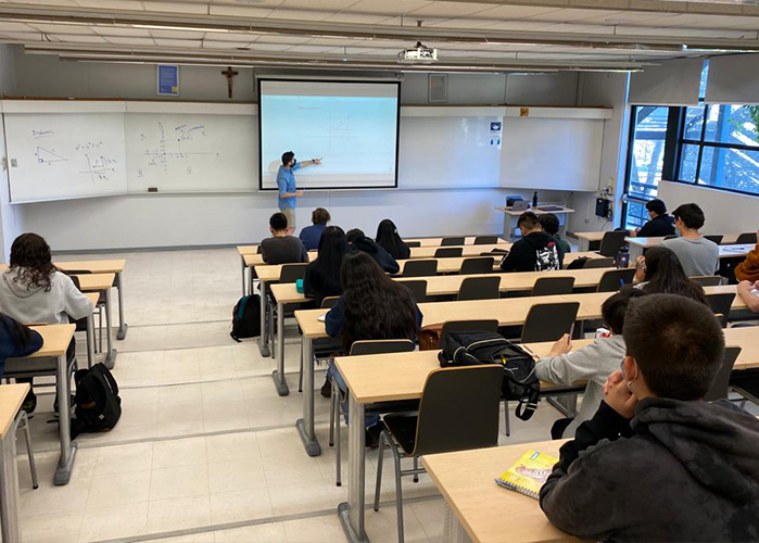 Profesor mirando y apuntando con su mando izquierda datos en una presentación proyectada en pizarrón, frente a estudiantes sentados en sala de clases, se les ven sólo las espaldas.