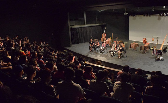 Fotografía de un teatro. En el escenario se ven cuatro personas sentadas, con instrumentos.