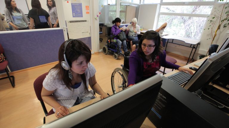 Dos estudiantes que participan del Programa para la Inclusión de Alumnos con Necesidades Especiales en las instalaciones del PIANE UC, sentadas en escritorio adaptado para la movilidad de estudiantes en silla de ruedas. Sobre el escritorio, computadores con softwares de apoyo a estudiantes con discapacidad.