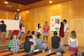 La imagen muestra a un grupo de cuatro estudiantes presentando un “poster” sobre la salud, en el escenario de un auditorio. En la parte inferior, diez personas escuchan atentamente la exposición.