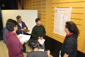 La imagen muestra a dos estudiantes presentando un poster sobre el método Cornell de apuntes, alrededor de ellos, cuatro personas evalúan sobre algunos papeles la presentación.