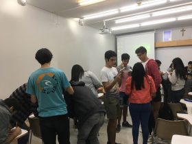 En la imagen se observa una sala de clases de la UC con estudiantes que se encuentran de pie con papeles en sus manos, algunos de ellos escribiendo en sus documentos.