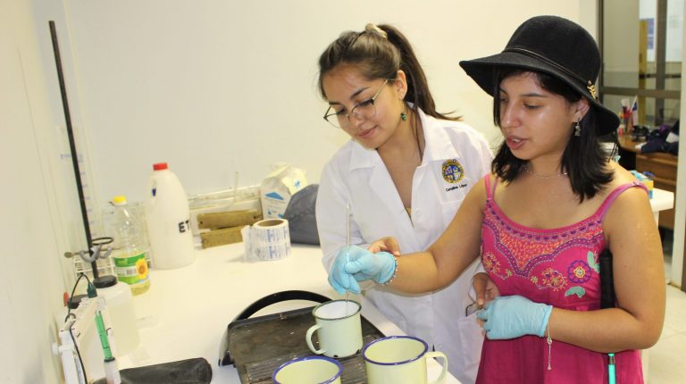 En la foto estudiante ciega es guiada por ayudante en Laboratorio de Química hacia una plancha con vasos para un experimento.