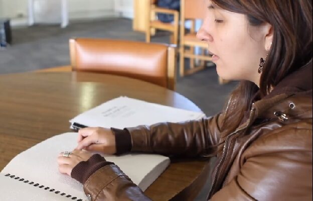 Estudiante ciega leyendo documento en Braille, sentada en mesa de la Biblioteca San Joaquín.