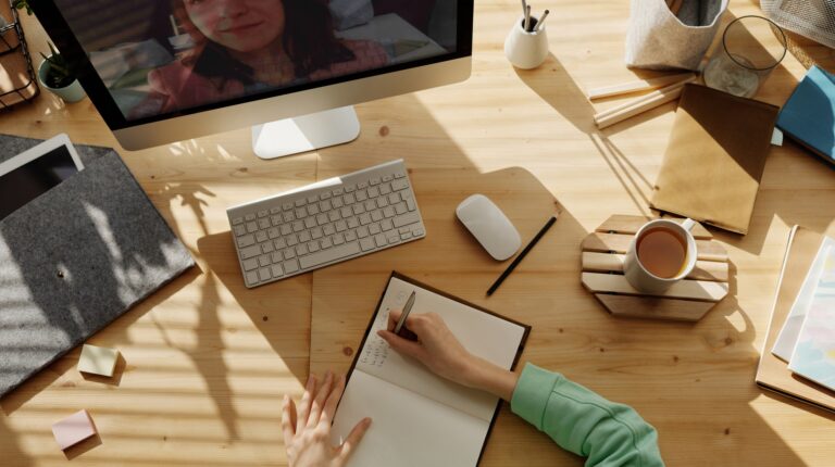 Se ve una mesa desde arriba, una persona tomando apuntes con elementos de escritorio a su alrededor y viendo una clase online.