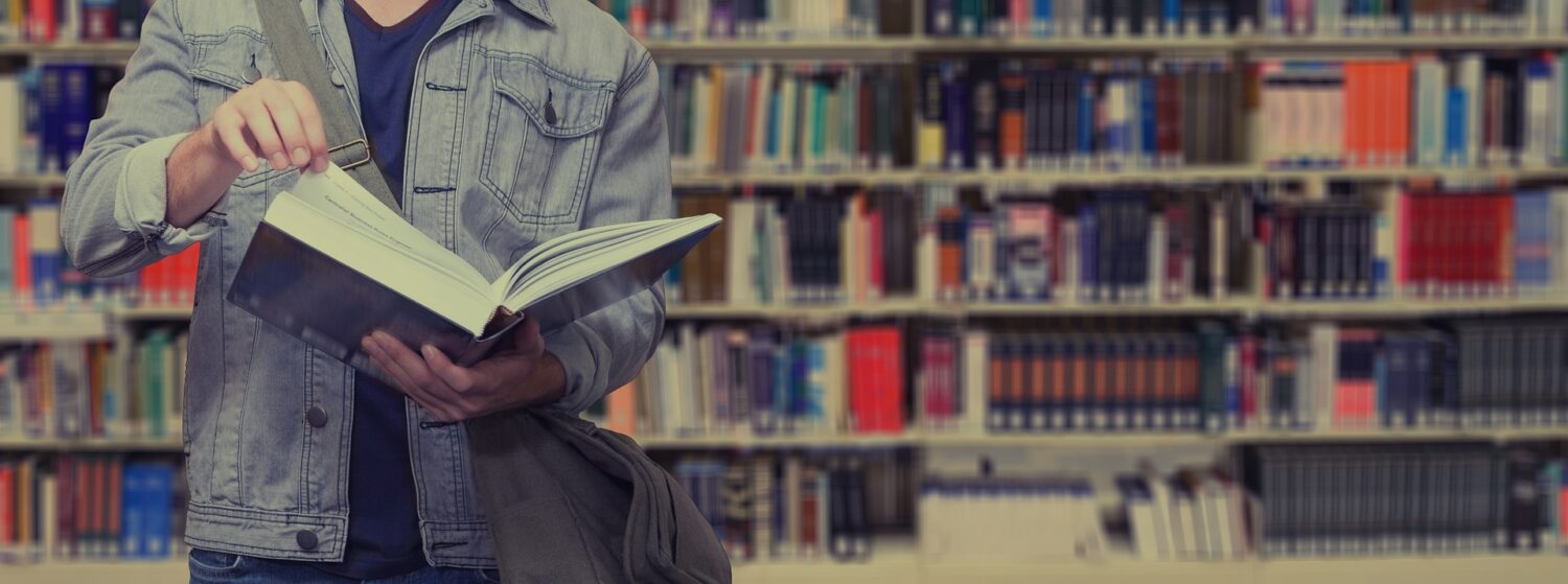 Torzo de hombre con camisa gris, leyendo un libro grande. De fondo se ve una biblioteca llena de libros.