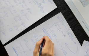 Se ve la mano de una persona escribiendo ejercicios matemáticos sobre papeles con fórmulas escritas a mano.