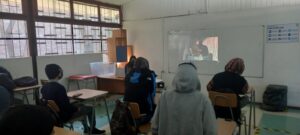 Estudiantes escolares de liceos PACE UC en actividad presencial en colegio, específicamente de espaldamirando el pizarrón donde se encuentra proyectada una película.