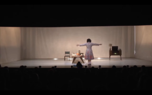 Se ve un escenario, una actriz de espaldas, en la escena de una obra de teatro. En la escena se visualiza una habitación con una mesa llena de pequeños objetos, un mueble pequeño y una silla.