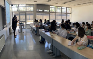 Profesora de pie, de perfil, haciendo gesto con sus manos frente a estudiantes sentados en sala de clases mirándola. El contexto de la clase es taller de nivelación académica.