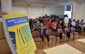 Folleto con el nombre "Alertas Académicas UC" impreso en la portada de una libreta. De fondo de manera borrosa se perciben personas sentadas conversando en un auditorio.