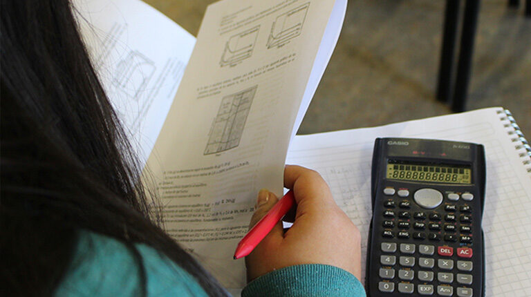 Foto de estudiante de espalda, leyendo una guía, de fondo se visualiza sobre la mesa de su asiento un cuaderno y una calculadora.
