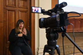 Fotografía de una intérprete en lengua de señas haciendo una seña frente a una cámara de video que la está grabando
