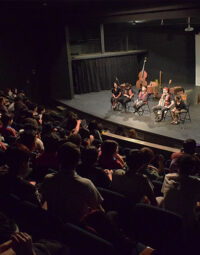 Fotografía de un teatro. En el escenario se ven cuatro personas sentadas, con instrumentos.