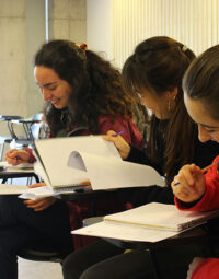 Tres jóvenes sentadas sonriendo mirando y escribiendo en cuadernos, se encuentran sentadas en una sala de clases