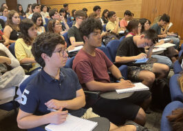 Foto de decenas de estudiantes sentados con papeles sobre a mesa de los asientos, mirando todos hacia el mismo lugar