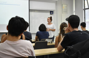 Foto de profesora UC en sala de clases frente a estudiantes sentados en la sala