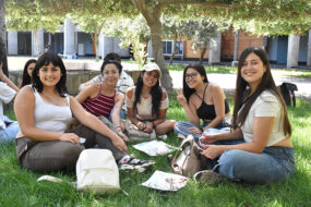 Foto de grupo de estudiantes UC sentadas en el pasto mirando a la cámara y sonriendo