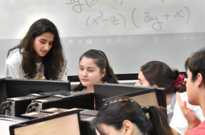 Foto de estudiantes frente a computadores, junto a otra persona de pie señalando uno de los computadores