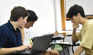 Foto de tres estudiantes sentados en una sala de clases conversando con papeles y aparatos electrónicos en sus manos