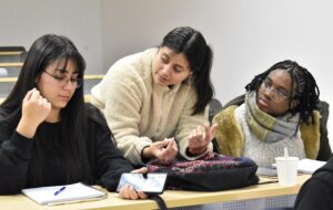 Fotografía de tres estudiantes conversando entre sí, mirándose y haciendo gestos con las manos sentadas en un mesón en una sala de clases