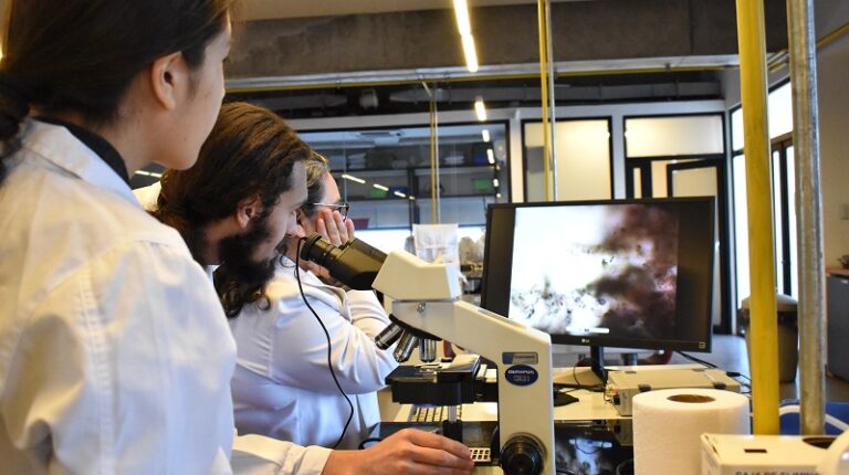 Fotografía de tres personas con bata blanca, una de ellas con sus manos en un microscopio, todas mirando una pantalla
