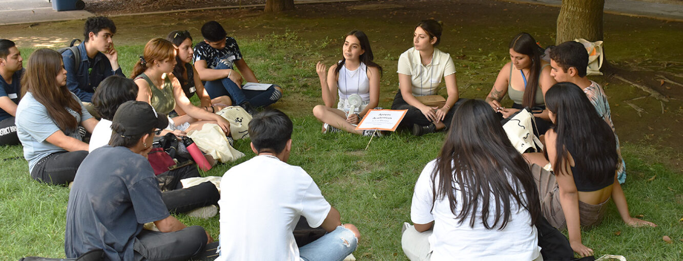 Grupo de estudiantes sentados en el pasto en torno a una estudiante a quién están mirando