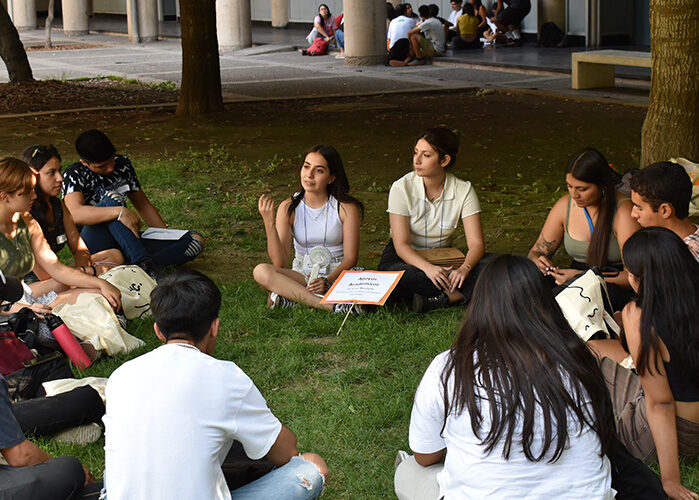 Grupo de estudiantes sentados en el pasto en torno a una estudiante a quién están mirando