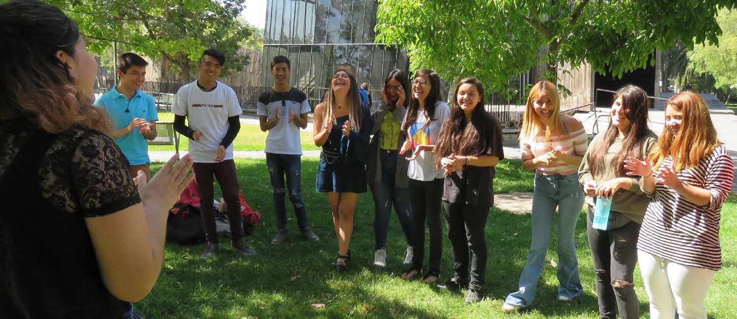Alumnos y alumnas de PACE UC sonriendo y aplaudiendo en actividad dirigida por tutora del programa, realizada en el patio exterior del edificio Cddoc en Campus San Joaquín.