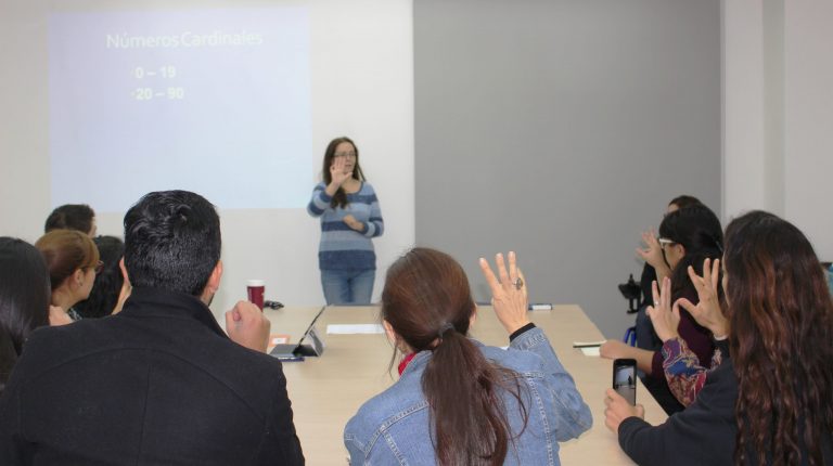 Profesores y profesoras en clase de Lengua de Señas Chilena haciendo una seña con su mano derecha que muestra la profesora al final de la sala.