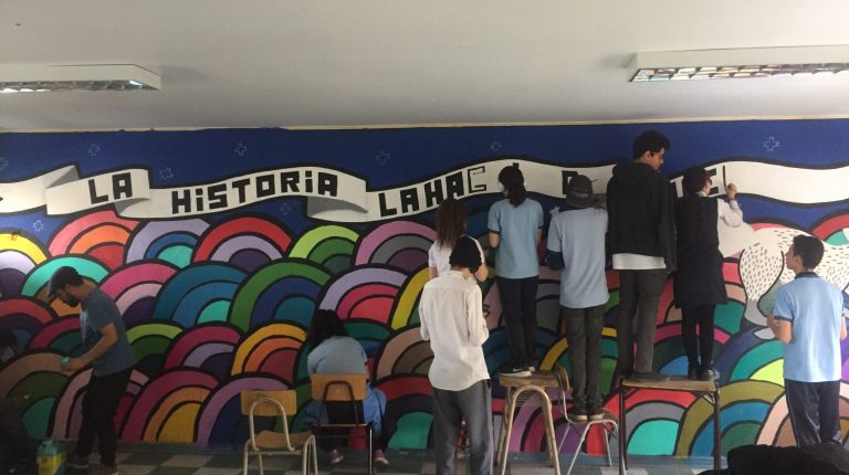 Siete estudiantes junto al encargado del taller Claudio Gacitúa, de espaldas a la cámara pintando un mural en la sala de clases de Historia del Liceo Gabriel González Videla A24 con la frase “La historia la hacen los pueblos”.