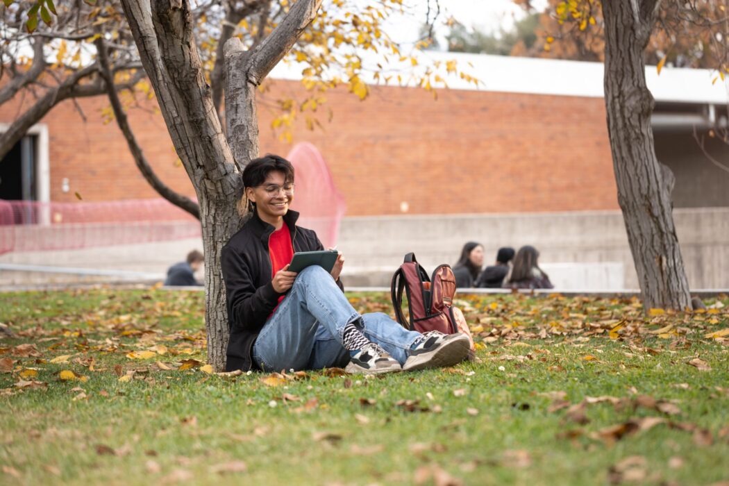 Estudiante sentado en el césped bajo un árbol, leyendo en una tablet y sonriendo